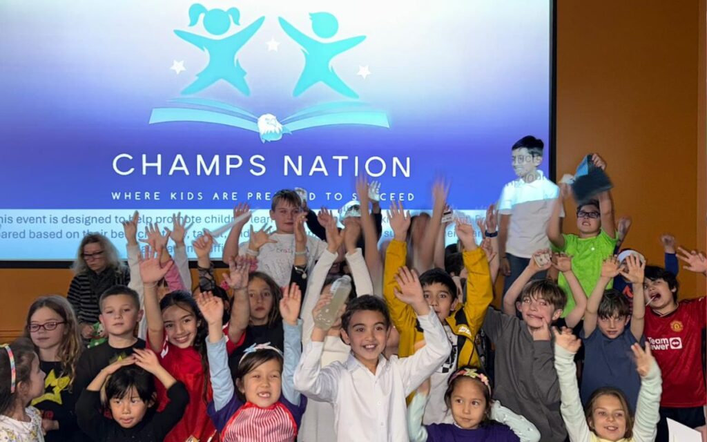 Group of smiling children with hands raised in a classroom or event space a large CHAMPS NATION banner on screen behind them