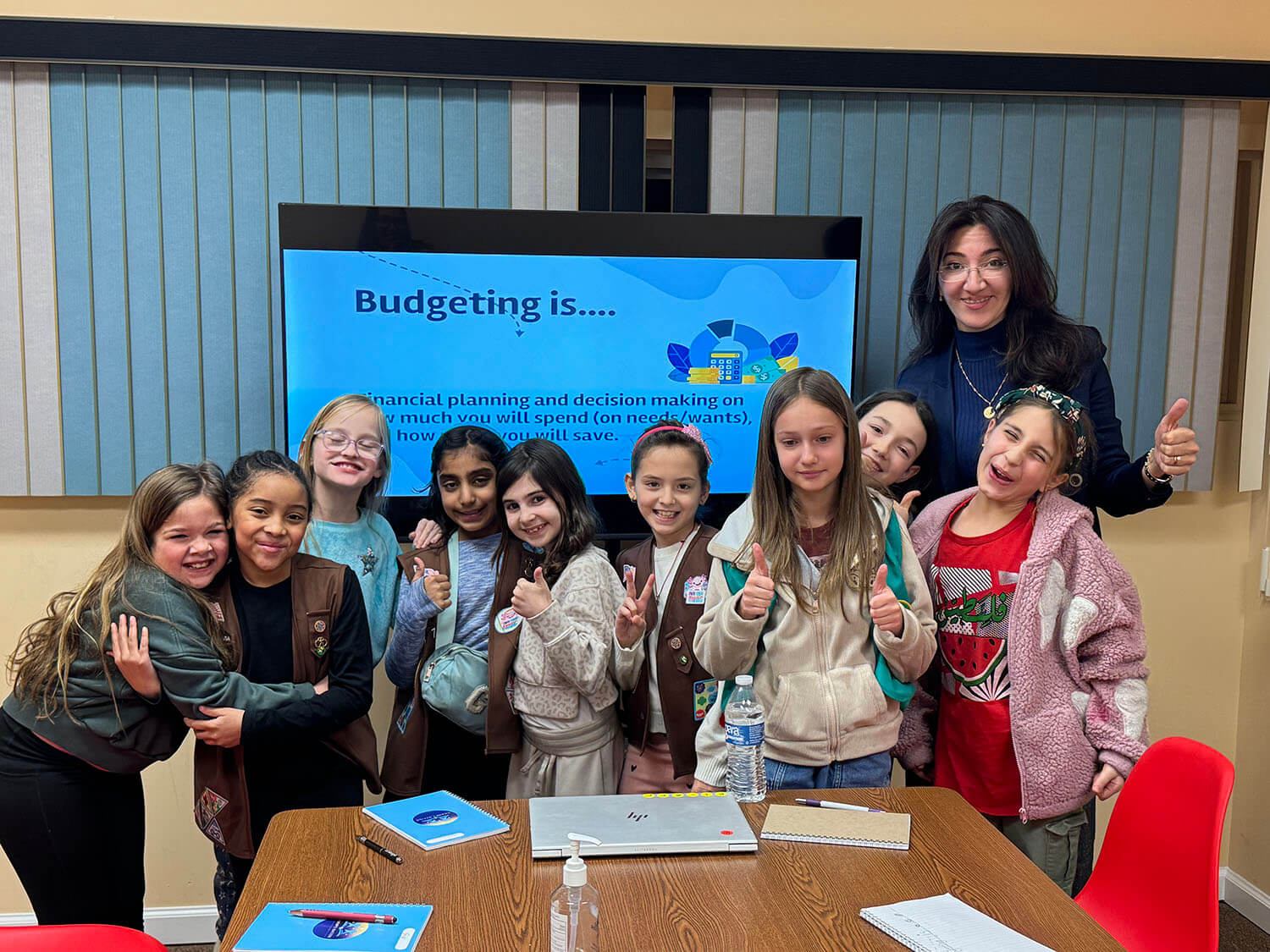 Group of smiling kids and a female teacher posing in a classroom in front of a screen that reads 'Budgeting is...'.