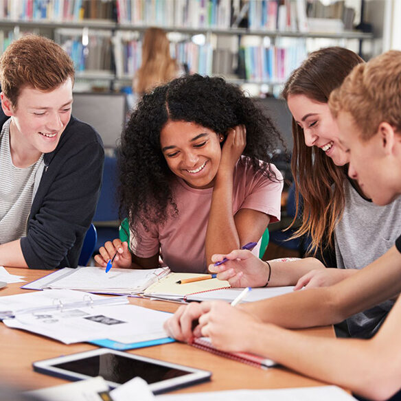 Group of four students studying together at a library table smiling and discussing papers